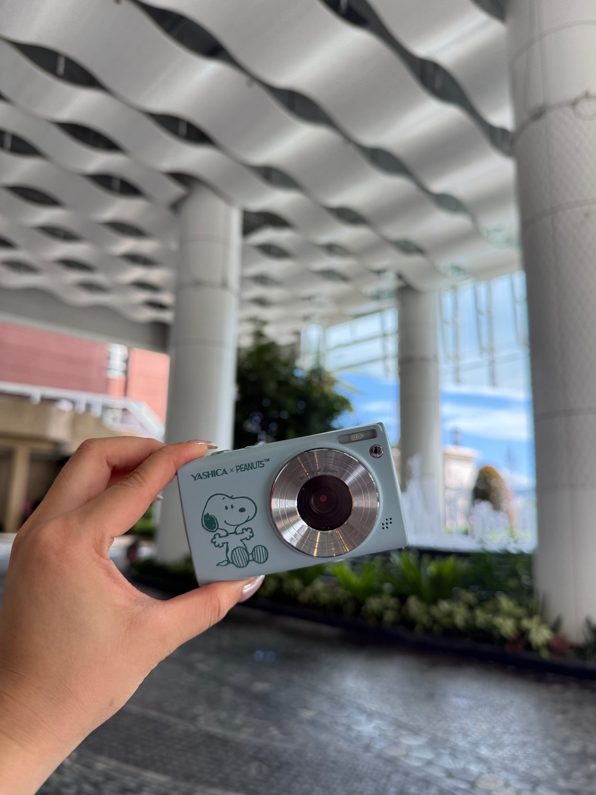 Hand holding a small metallic digicam with a textured surface and circular design, outdoors.